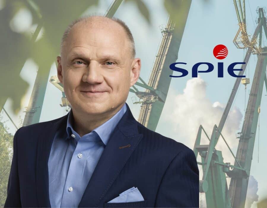 A man in a suit stands in front of industrial cranes and the SPIE logo on a blue sky background.