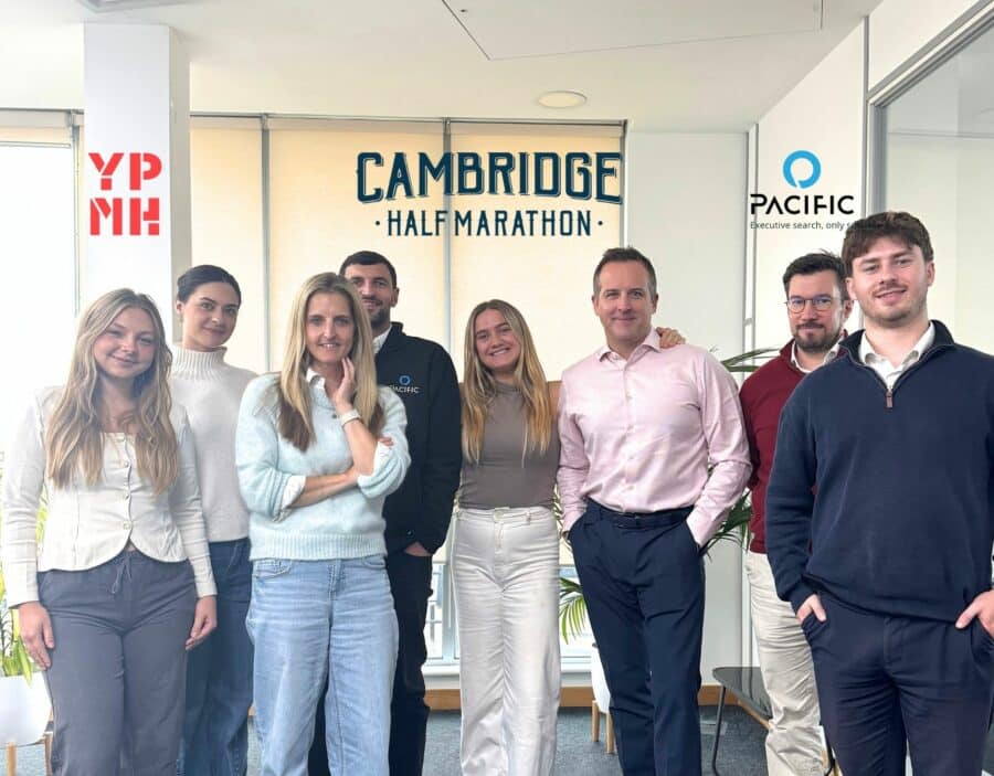 A group of eight people stand together in an office with the Cambridge Half Marathon logo on the window behind them.